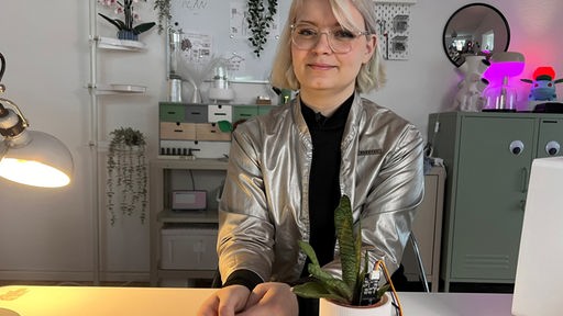 Frau sitzt am Schreibtisch und hat Roboterpflanze vor sich. Im Hintergrund sind mehr Roboter zu sehen