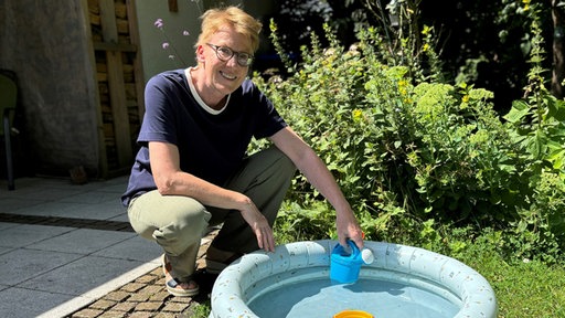 Frau hockt an einem Kinderpool und hält kleine Gießkanne in der Hand