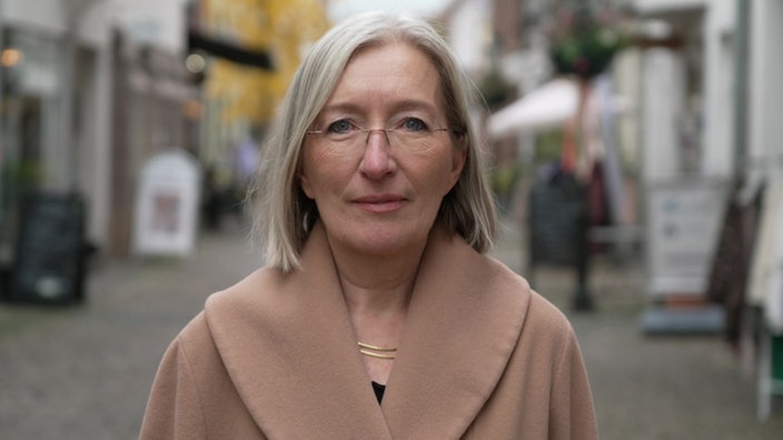 Eine Frau mit rahmenloser Brille steht in der Mitte einer leeren Einkaufsstraße und schaut in die Kamera.