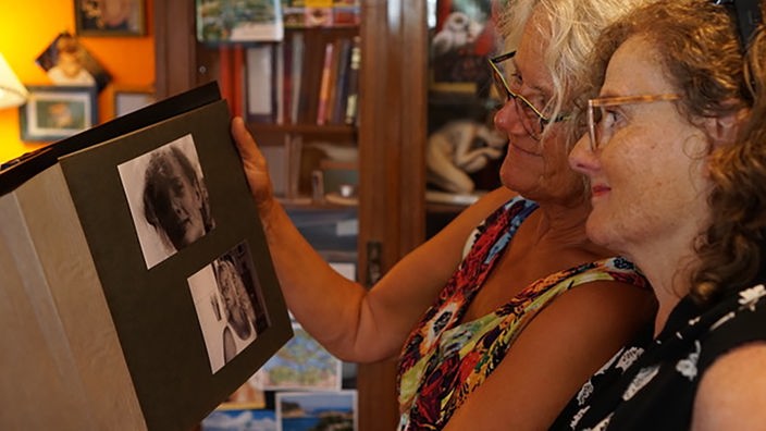 Zwei ältere Frauen stehen zusammen und schauen sich gemeinsam ein Fotoalbum an. Auf der aufgeschlagenen Seite sind zwei Bilder von den beiden zu sehen