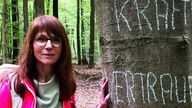 Frau mit Brille berührt ein Baum im Wald und schaut freundlich zur Kamera