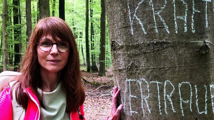 Frau mit Brille berührt ein Baum im Wald und schaut freundlich zur Kamera