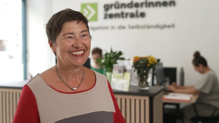 Eine lächelnde Frau steht in einem Büro vor der Rezeption. Im Hintergrund ist das Logo der Gründerinnenzentrale zu sehen. Zwei weitere Frauen sitzen am Schreibtisch und arbeiten am Computer.