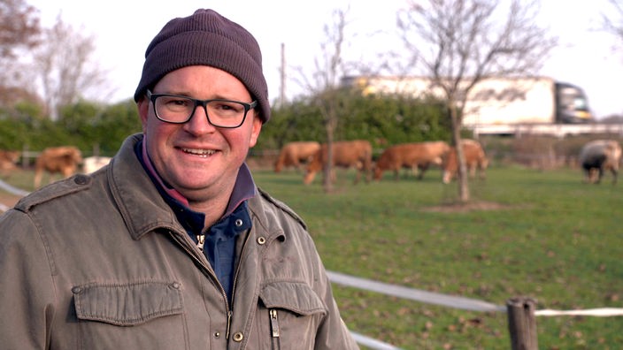 Ein Mann schaut lächelnd in die Kamera, im Hintergrund eine Weide mit Kühen und ein Lastwagen
