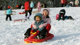 Zwei Kinder rodeln auf ihrem Schlitten einen verschneiten Hang hinunter
