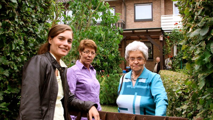 Zwei ältere Frauen stehen mit einer jüngeren Frau am Gratenzaun, im Hintergrund ein Reihenhaus