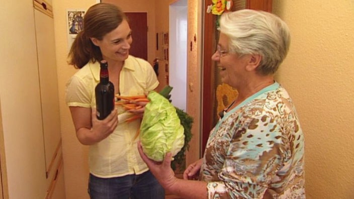 Eine junge Frau steht mit einer älteren Frau im Hausflur, beide haben Einkäufe im Arm