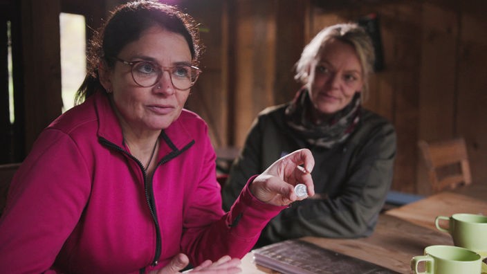 Zwei Frauen sitzen in einer Waldhütte, eine hält ein 50-Pfennig-Stück zur Kamera