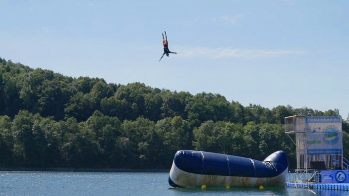 Blick auf einen See, auf dem ein großes Luftkissen schwimmt, von dem gerade ein Mensch in die Luft gesprungen ist