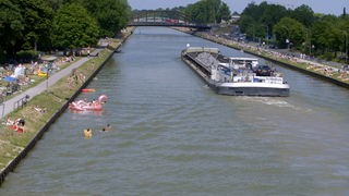 Ein Binnenschiff auf dem Kanal, am Ufer liegen Menschen, im Wasser sind Badende
