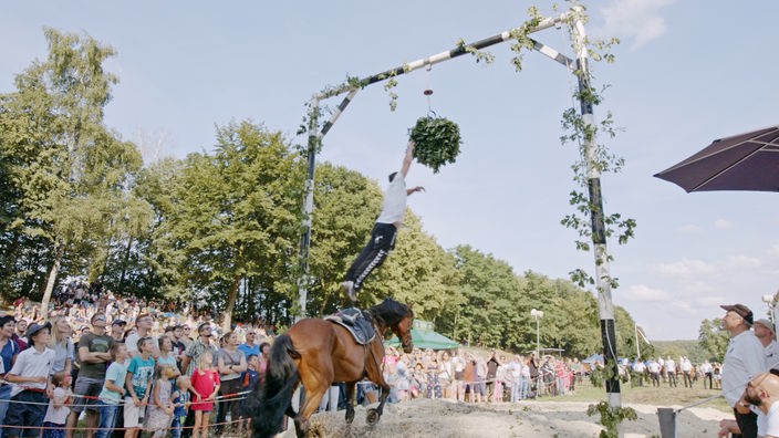 Ein Mann springt von einem Pferd in die Höhe und versucht, einen aufgehängten Kranz herunterzureißen