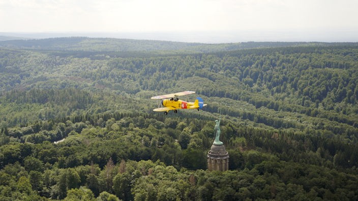 Ein gelber Doppeldecker fliegt über den Teutoburger Wald am Hermannsdenkmal vorbei