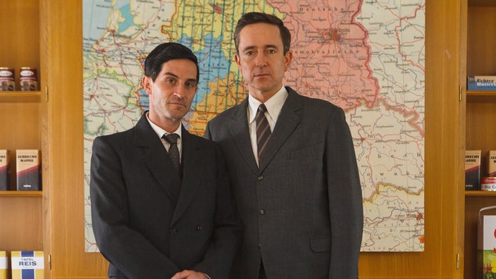 Porträtbild der beiden Schauspieler, die die Aldi-Brüder spielen, in einem Büro vor einer Deutschland-Karte