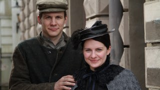 Szenenbild: Ein junger Mann und eine junge Frau in Kleidung um 1900 schauen lächelnd in die Kamera
