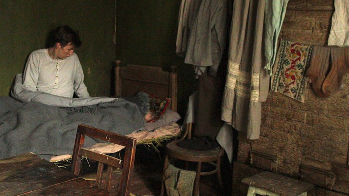 Szenenbild: Ein junger Mann liegt in einer Wohnküche im Bett 