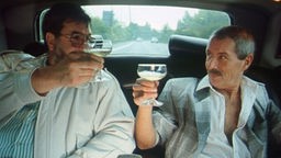 Zwei Männer sitzen im Fond eines Autos und prosten sich mit Champagnergläsern zu