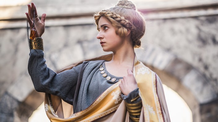 Spielszene: Eine Frau in antiker römischer Tracht betrachtet einen Bernstein-Anhänger