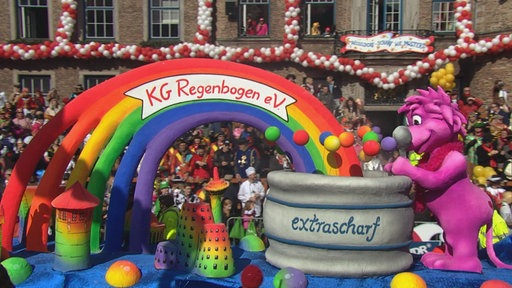 Der Rosenmontagszug zieht am Düsseldorfer Rathaus vorbei, im Vordergrund ein Mottowagen mit einem pinken Löwen an einem Senftopf, auf dem extrascharf steht