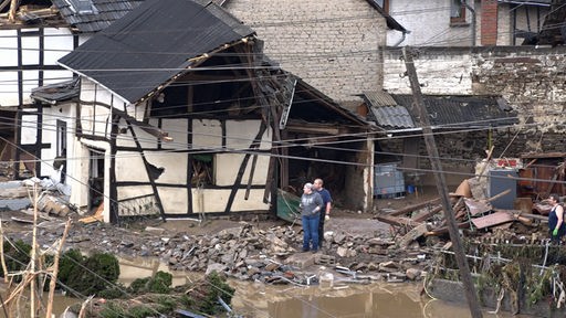 Blick auf ein zerstörtes Haus, ein Mann und eine Frau stehen davor im Schlamm