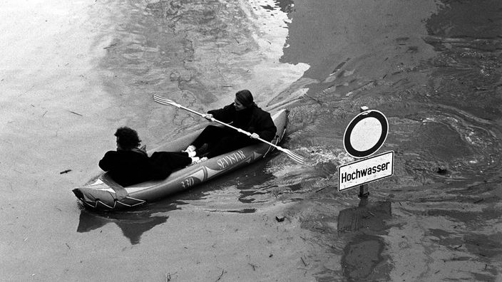 sw-Foto: zwei junge Menschen paddeln in einem Schlauchboot an einem Hochwasser-Schild vorbei.