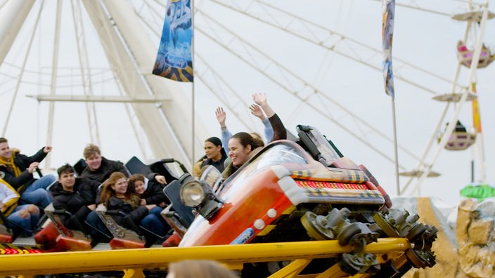 Menschen fahren lachend in einer Achterbahn, im Hintergrund ein Riesenrad