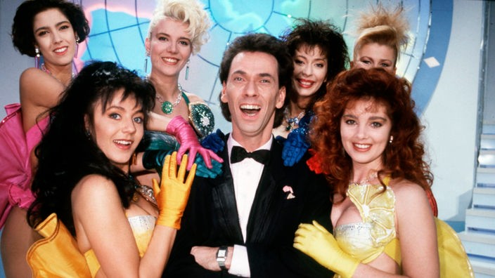Hugo Egon Balder im Smoking steht in der Kulisse der Tutti Frutti Show, umgeben von leichtbekleideten Frauen.