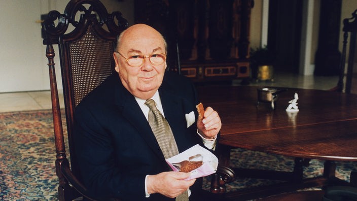 Ein älterer Mann sitz am Tisch, eine Tafel Schokolade in der einen und einen Riegel Schokolade in der anderen Hand