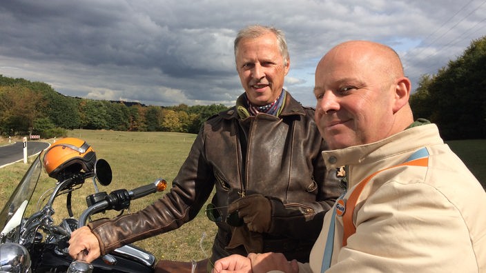 Zwei Männer stehen an ihren Motorrädern, im Hintergrund waldige Landschaft