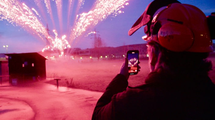 Ein Mann filmt auf einem Versuchsgelände mit einem Smartphone ein Feuerwerk.