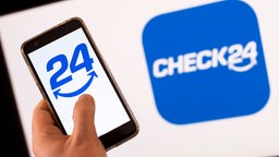 Ein Iphone und mit dem Check24-Symbol wird vor einem Bildschirm hochgehalten. 