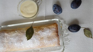 Ein Pflaumenstrudel auf einer Porzellan-Platte mit Pflaumen zur Deko und einem Schälchen Vanillesauce