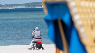 Eine ältere Frau sitzt auf ihrem Rollator hinter einem Strandkorb am Wasser.