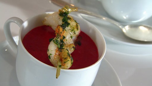 Rote Bete Suppe in einer Suppentasse angerichtet, darübergelegt ein Spieß mit Garnelen 