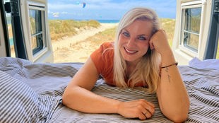 Frau mit langen, glatten blonden Haaren, blauen Augen und orangem T-shirt lehnt sich mit Blick in die Kamera auf ein Bett im Campervan mit schwarz-weiß gestreifter Bettwäsche, im Hintergrund Dünen, Meer, blauer Himmel, Kitesurf-Segel