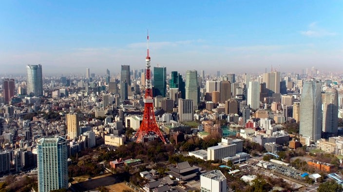Blick von schräg oben auf eine riesige Großstadt mit vielen Wolkenkratzern, in der Mitte ein rot-weißer nach oben schmaler werdender Turm in Stahlfachwerkbauweise