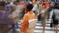 im Vordergrund eine dunkelhaarige Frau in lachsfarbenem Kimono mit dem Handy telefonierend von hinten, um sie herum viele unscharf zu sehende Fußgänger auf einem Zebrastreifen  