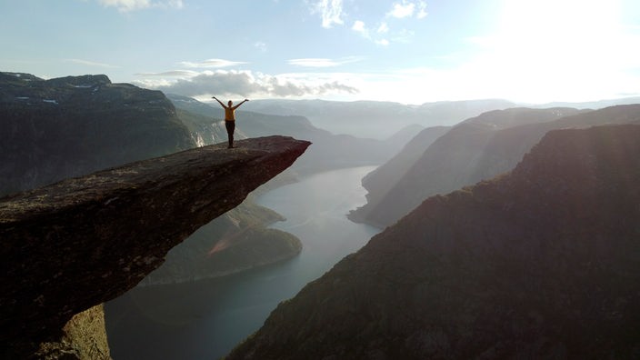 eine Person im Gegenlicht auf einer Felszunge mit Blick auf eine Fjordlanschaft mit steilen Bergen zu beiden Seiten des Wassers