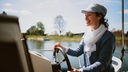 eine fröhlich lachende Frau mit Schlägerkappe am Steuer eines Bootes, im Hintergrund Wasser und blauer Himmel