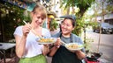 zwei Frauen, eine mit dunklen, eine mit hellen Haaren, stehen lachend auf einem Gehweg und halten jeweils einen Teller mit einem Nudelgericht und Essstäbchen in der Hand