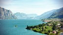 türkisblauer großer See zwischen felsigen Bergen, am rechten Ufer einige Steinhäuser und weiter hinten ein Dorf zwischen üppig grüner Vegetation