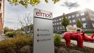 Ein Wegweiser mit dem Elmos-Logo steht vor dem Firmengebäude und daneben ein rotes Dortmunder Nashorn mit dem Elmos-Logo.