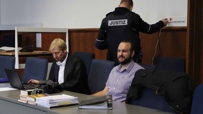 Der Angeklagte sitzt in kariertem Hemd an einem grauen Tisch vor Aktenordnern. Links sitzt sein Straverteidiger, im Hintergrund steht ein Justizbeamter.