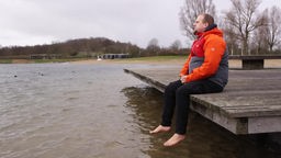 Alexander Tok sitzt auf einem Steg am Wasser und baumelt mit den nackten Füßen