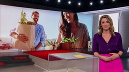 Die Moderatorin Catherine Vogel steht im Markt-Fernsehstudio, Im Hintergrund sieht man zwei Personen, die Einkaufstüten tragen.