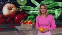 Catherine Vogel steht im Markt.Fernsehtstudio und hält eine Orange in ihren Händen. Hinter ihr sieht man einen Obst-und Gemüsekorb.