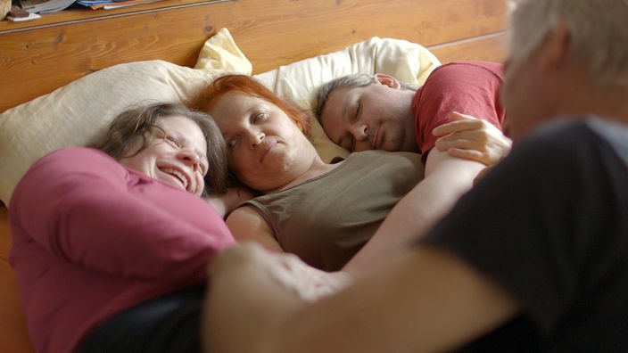 Eine Frau liegt mit zwei Männern an ihrer Seite in einem Bett. Über sie gebeugt ist ein weiterer Mann.