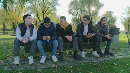 Fünf männliche Jugendliche sitzen auf einer Parkbank. Drei von ihnen halten Smartphones in der Hand.