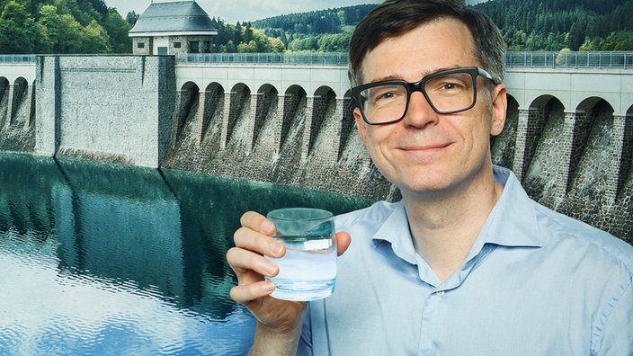 Ralph Caspers vor einer Staumauer mit einem Glas Wasser in der Hand