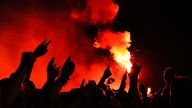 Zuschauer strecken ihre Hände nach oben in den von Pyrotechnik rot gefärbten Nachthimmel