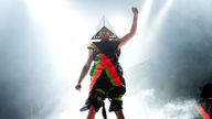 Ein Mitglied von Deichkind streckt den Arm in die Luft. Er trägt ein Kostüm aus unterschiedlichen Stofffetzen in Neonfarben 
